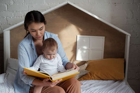 Loving young mother sitting in cozy kids room with curious baby daughter on lap reading fairy-tale...