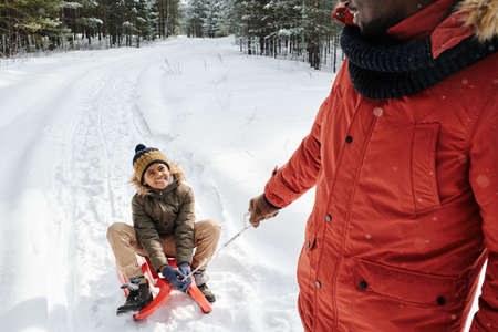 Happy cute little boy in winterwear looking at his father pulling sledge with him while moving along...
