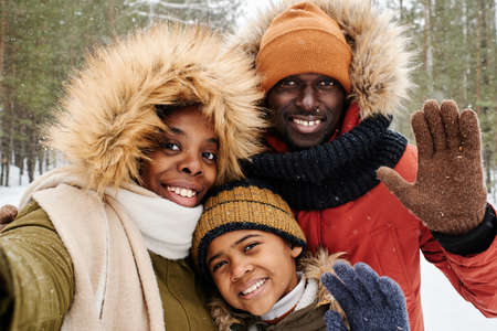 African American family of three in winterwear greeting you while looking at camera in park or forest on frosty winter day at leisureの写真素材