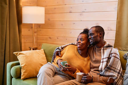 Happy young restful couple with cups of hot tea watching television in living room while relaxing on...