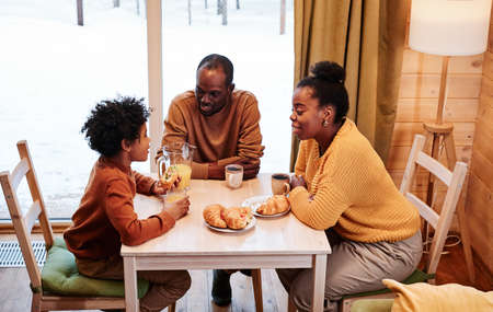 Young happy family of three communicating by breakfast while sitting by table served with homemade...