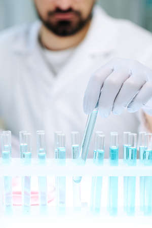 Gloved hand of young contemporary male scientist taking one of several flasks containing blue liquid...