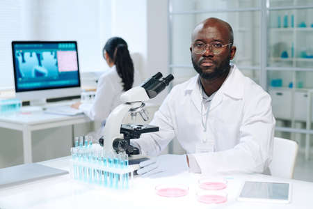 Young serious scientist in lab coat sitting by workplace in front of microscope and looking at...
