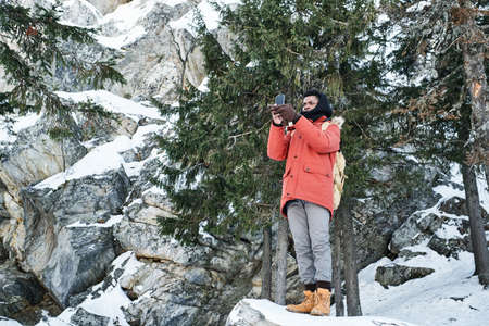 Long shot of young Black man hiking in mountains on winter day standing on rock taking pictures on...
