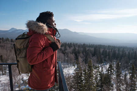 Young African American guy wearing backpack standing at observation deck on mountain top on winter...