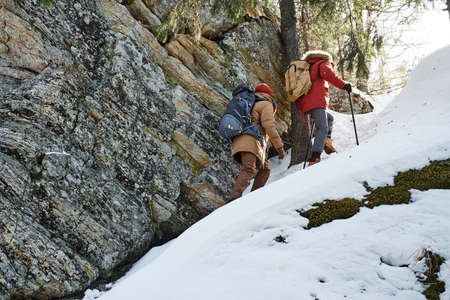 Two unrecognizable young men wearing backpacks hiking together in mountains on winter day, low angle...