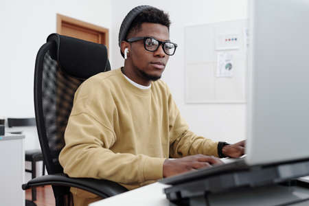 Young contemporary serious male it programmer in yellow sweatshirt looking at computer screen while...