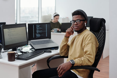 Young confident black man in casualwear with earphones sitting by workplace with compute monitor and laptop with decoded dataの写真素材