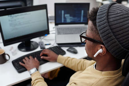 Above angle of young black man in casualwear listening to music in earphones while decoding data in...