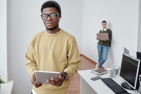 Contemporary young male programmer with tablet looking at camera against female coworker with laptop...