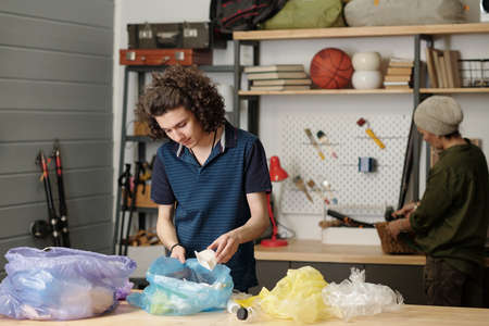 Youthful guy in casualwear putting plastic food containers into one of several cellophane sacks...