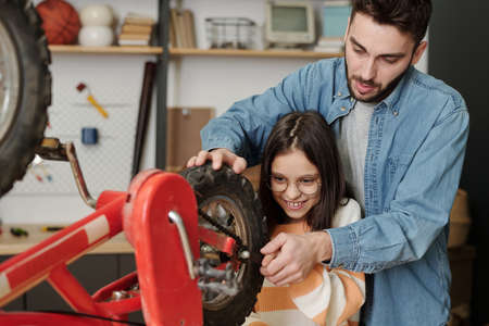 Smiling little girl helping father repair her bicycle while holding part of wheel and using wrench...