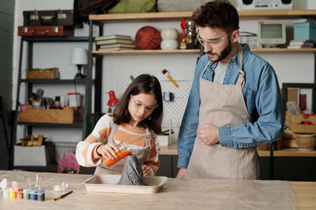 Father and daughter in aprons and protective eyeglasses pouring orange liquid into crater of volcano...