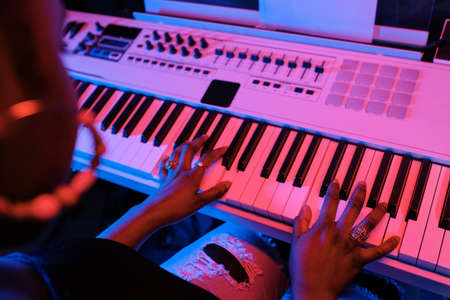 High angle view of unrecognizable young Black woman playing synthesizer in recording studio in neon...