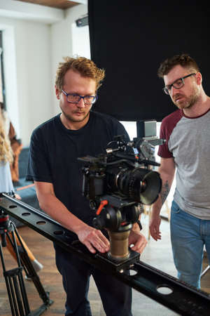 Young professional videographer moving video camera along slider while adjusting its options for...