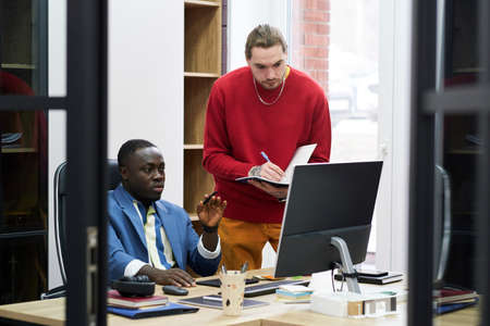 African manager sitting at his workplace with computer and giving work to businessman while he...