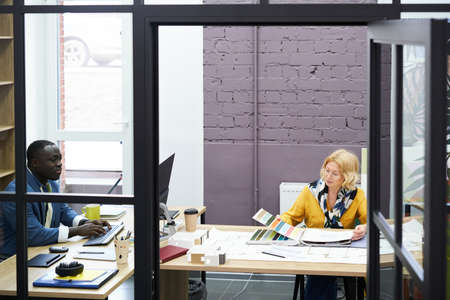 Designers sitting at table at modern office working on computer and with colour palette for future projectの写真素材