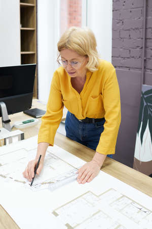 Serious female engineer in eyeglasses working with blueprint on table using pencil and rulerの写真素材