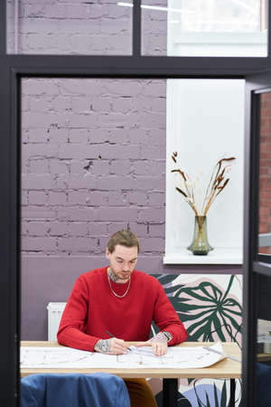 Young architect using ruler to draw blueprint of new construction project while sitting at workplace at officeの写真素材