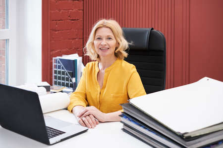 Portrait of mature businesswoman smiling at camera while sitting at desk with laptop and stack of...