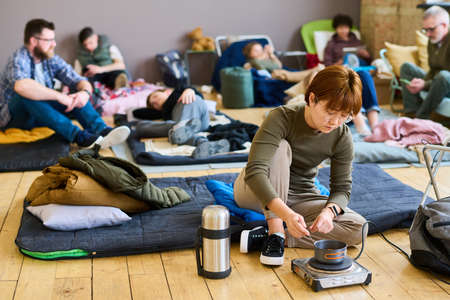 Young hungry Asian female refugee cooking food in small pan on electric stove on the floor while...