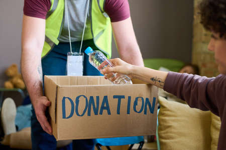 Young male volunteer in uniform holding donation box with food and drinks while female refugee...