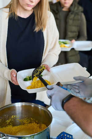 Young blond female refugee holding plastic container over pan with pilaf while male volunteer in gloves putting her prepared foodの写真素材
