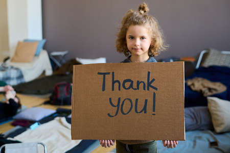Cute blond little boy with cardboard poster saying thank you looking at camera while standing against sleeping places in refugee campの写真素材