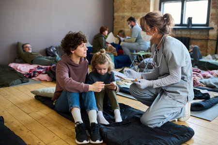Young female volunteer in medical uniform consulting refugee with child while sitting on squats on their sleeping place in frontの写真素材