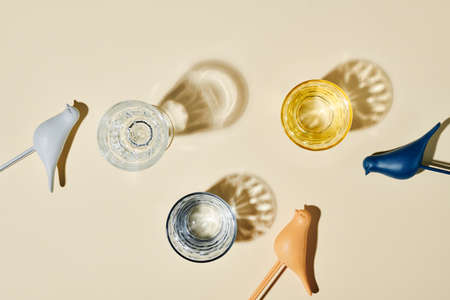 Flatlay of three glasses containing water, their shadows and small toy birds of various colors over...