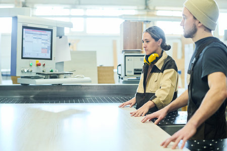 Two young workers in uniform working in team with wooden board on cutting machine at furniture...