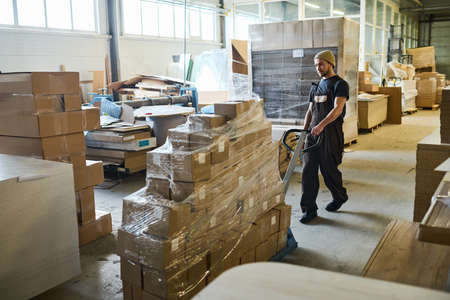 Young man in workwear carrying cart with stack of packed goods for transportationの写真素材