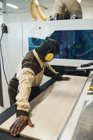 African worker in protective headset putting wooden plank on sawing machine to make furniture at...