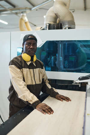 Portrait of African manual worker in uniform looking at camera while standing at modern equipment...