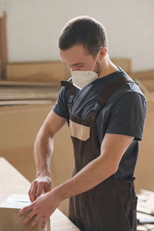 Young warehouse worker in protective mask packing boxes at table for further delivery