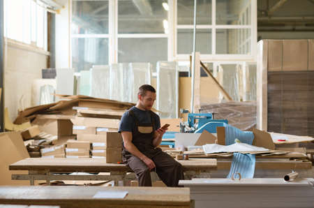 Manual worker sitting on table and using his mobile phone during his break after work in warehouse
