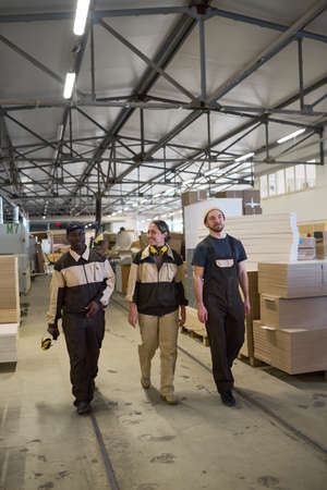 Group of workers in uniform walking along the corridor of warehouse with wooden materials in...