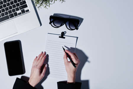 Overview of hands of businesswoman with pen over blank paper planning work by desk with laptop, smartphone and sunglassesの写真素材