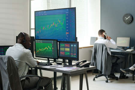 Modern African American and Caucasian men in formalwear sitting in front of computer monitors...