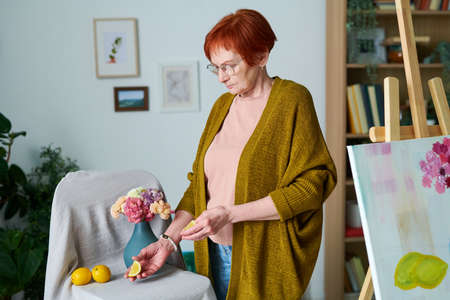 Elderly woman in eyeglasses making still life from lemons and flowers in vase before painting at...