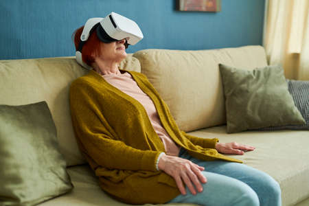 Senior woman in VR glasses using virtual reality for relaxation while sitting on sofa in living roomの写真素材