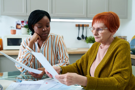 Senior woman in eyeglasses examining financial documents at table in kitchen together with social workerの写真素材