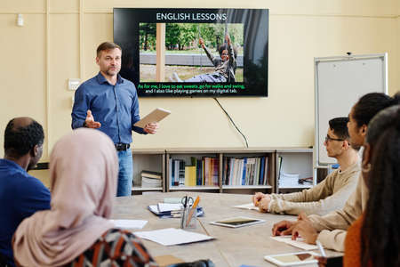 Professional English language teacher standing at table in front of multi-ethnic migrant students explaining something to themの写真素材
