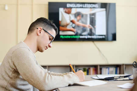 Serious young Middle Eastern man attending English language class writing something in notebook