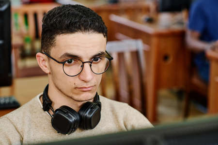 High angle close-up of serious Middle Eastern student wearing headphones working on computer in...