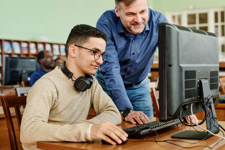 Cheerful mature teacher watching task his student finishing doing using desktop computer in libraryの写真素材