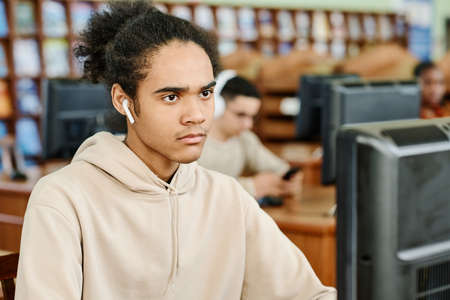 Young gen Z Black man wearing earphones sitting at desk in university library working on desktop...