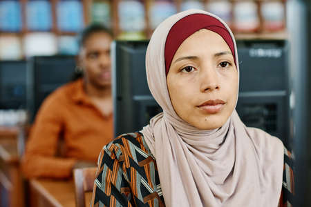 Portrait of young Muslim woman wearing hijab sitting in university library looking at computer...