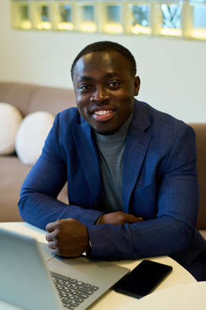 Portrait of African businessman in blue suit smiling at camera while sitting at table at restaurant with laptopの写真素材