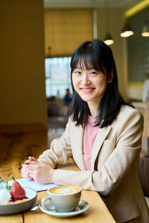 Portrait of ASian young businesswoman smiling at camera while sitting at table in cafe with dessert...